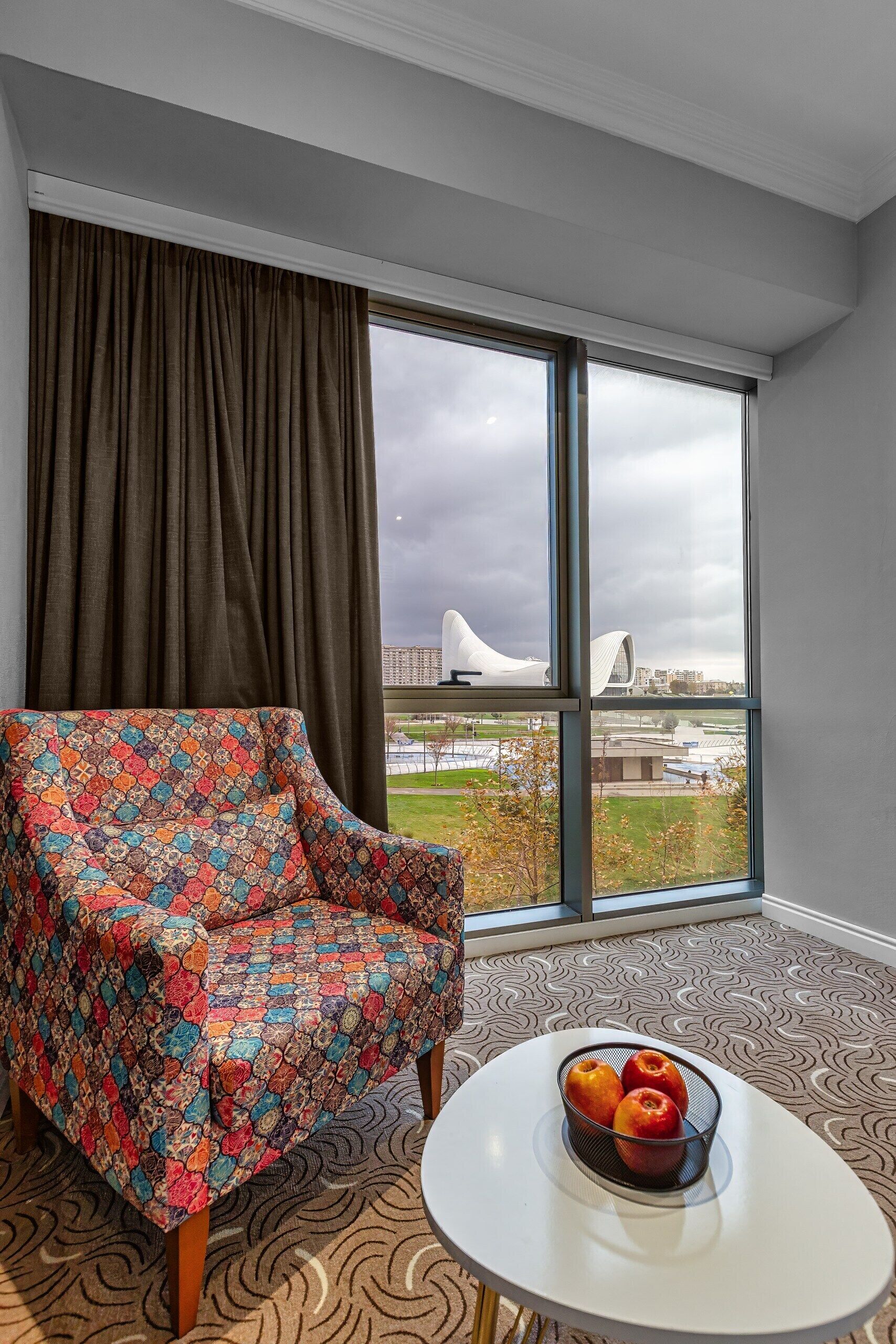 A room with a large window, a patterned armchair, and a table with a fruit bowl.