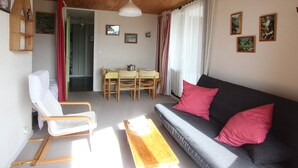 A cozy living room with a wooden ceiling, a grey sofa with red cushions, a wooden rocking chair, a dining table with chairs, and a wooden coffee table. - Comfortable ski-in/ski-out apartment with panoramic view (Chamrousse)