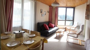 A cozy living room with a wooden ceiling, a sofa with red cushions, a dining table set with plates and glasses, and a window with a view of trees. - Comfortable ski-in/ski-out apartment with panoramic view (Chamrousse)
