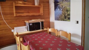 A small room with a wooden wall, a microwave, a table with a red tablecloth, and a painting of a mountain landscape.