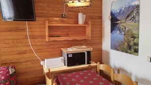 A room with wooden walls, a mounted television, a microwave, a table with a red tablecloth, and a painting of a mountainous landscape.