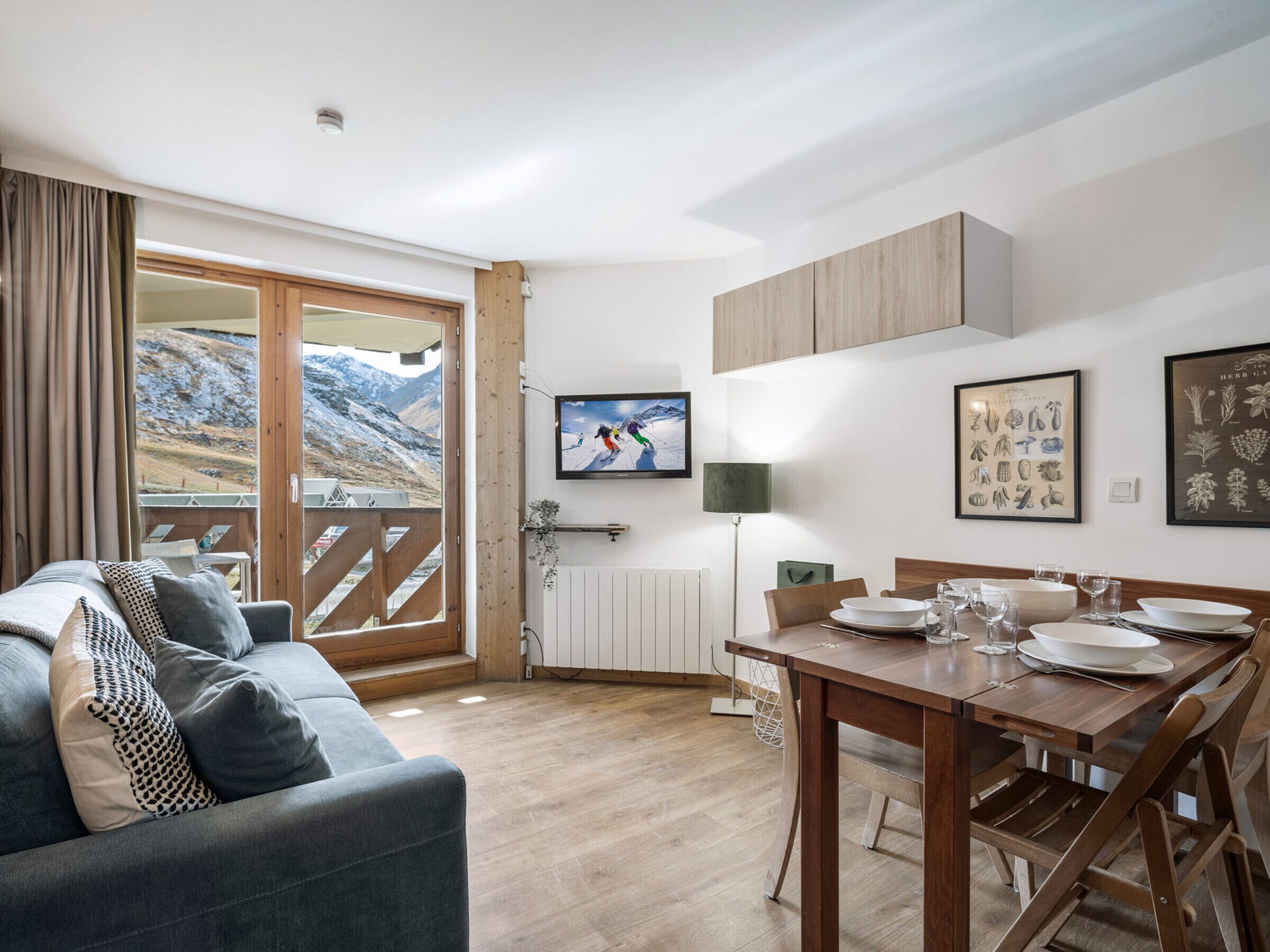 A modern living room with a sofa, a dining table set for four, and a large window with a view of mountains.