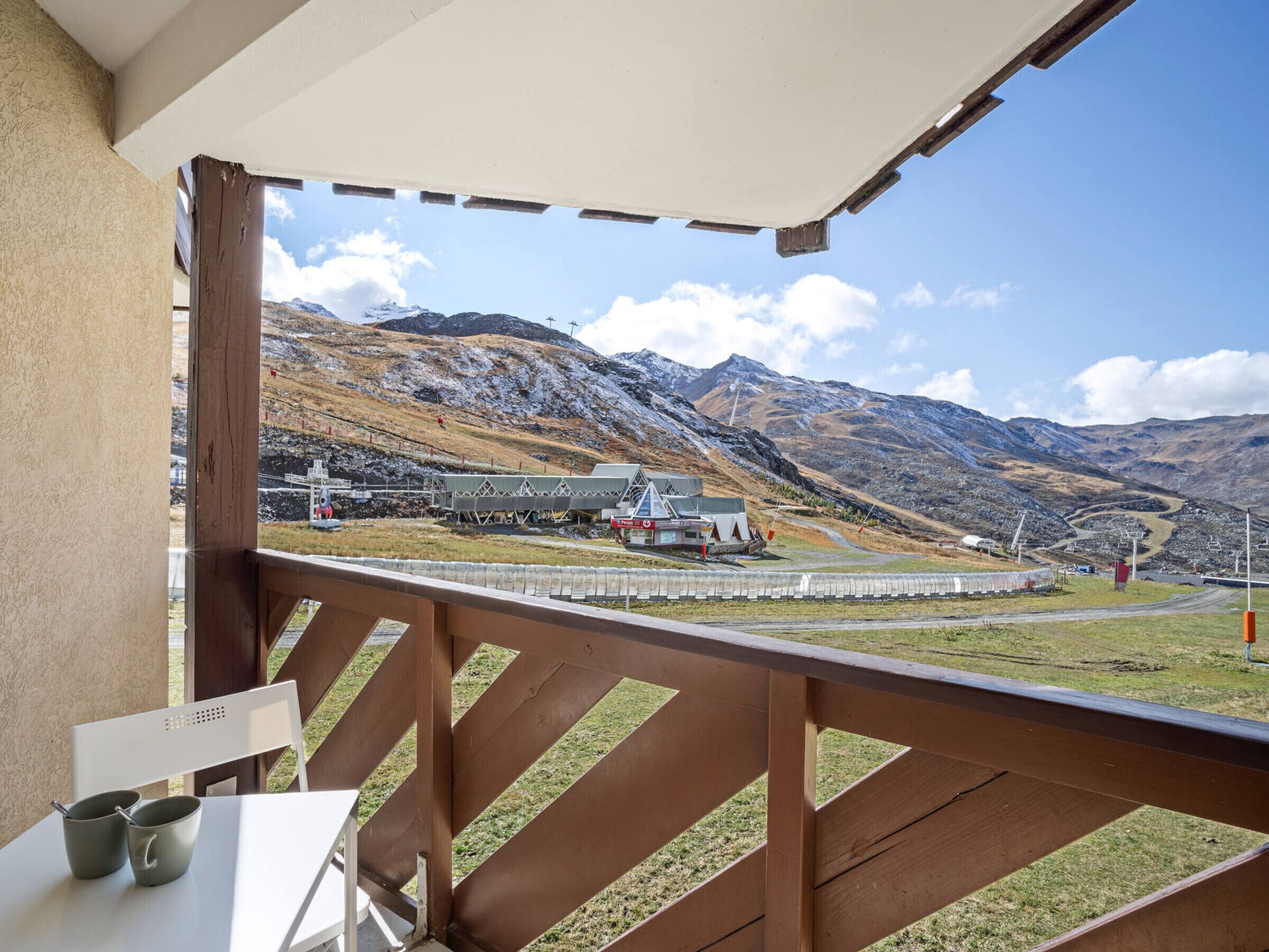 Un balcon donnant sur un paysage montagneux, un bâtiment avec un télésiège, et une table avec deux tasses.