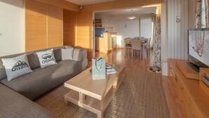 Ein modernes Wohnzimmer mit Sofa, Couchtisch und Fernseher. Im Hintergrund ist eine Küchenzeile mit Esstisch und Stühlen zu sehen.
