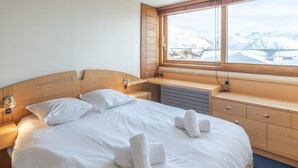 Ein Hotelzimmer mit Holz-Kopfteil, einem großen Bett mit weißen Bettwäsche, einem Fenster mit Blick auf schneebedeckte Berge und einer Holzkommode mit Schubladen.