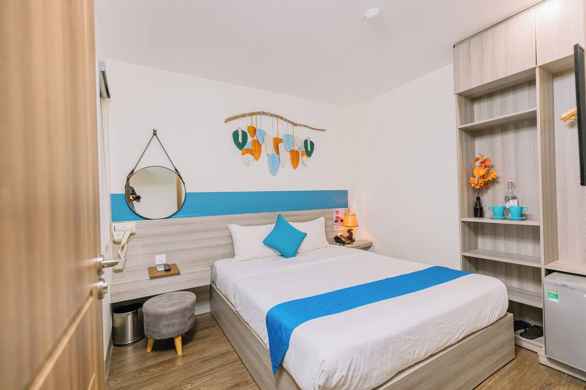A modern hotel room with a bed, a small desk, a mirror, a wall-mounted decoration, and a shelf with a bottle and a cup.