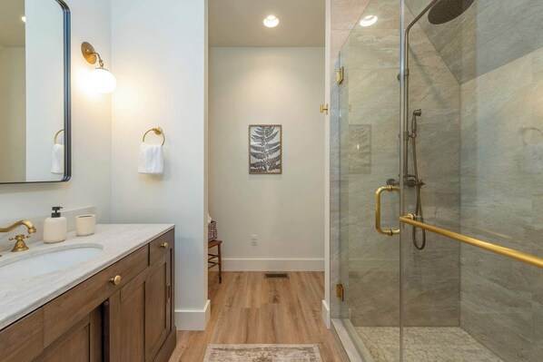 A bathroom with a marble sink, wooden cabinets, a glass shower enclosure, and a framed artwork on the wall.