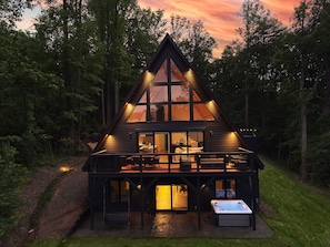 A modern, triangular wooden cabin with large windows, a hot tub, and a deck overlooking a forested area.