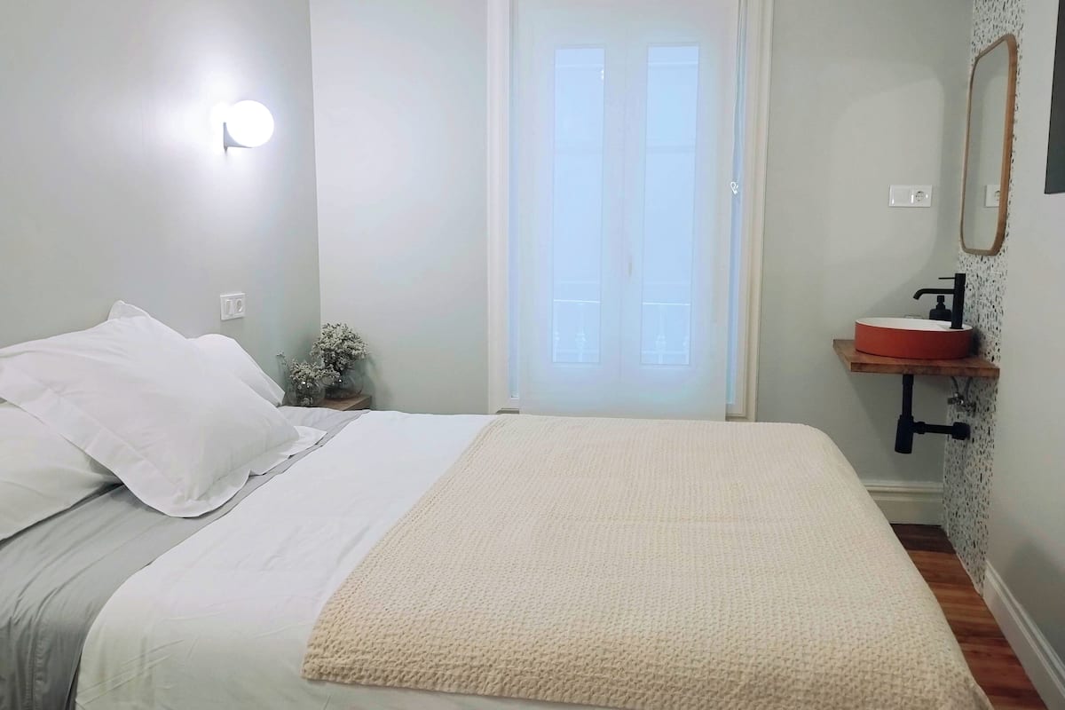 A bedroom with a bed, a bedside table with a vase, a window, and a bathroom with a sink and mirror.