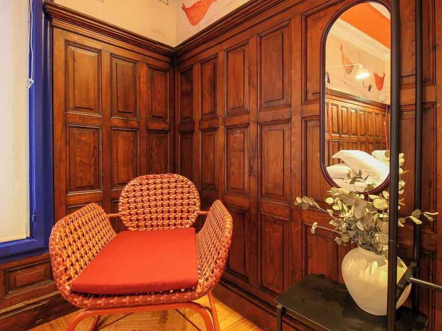 Habitación de esquina con paneles de madera, una silla de mimbre con cojín rojo y un espejo que refleja parte del cuarto.