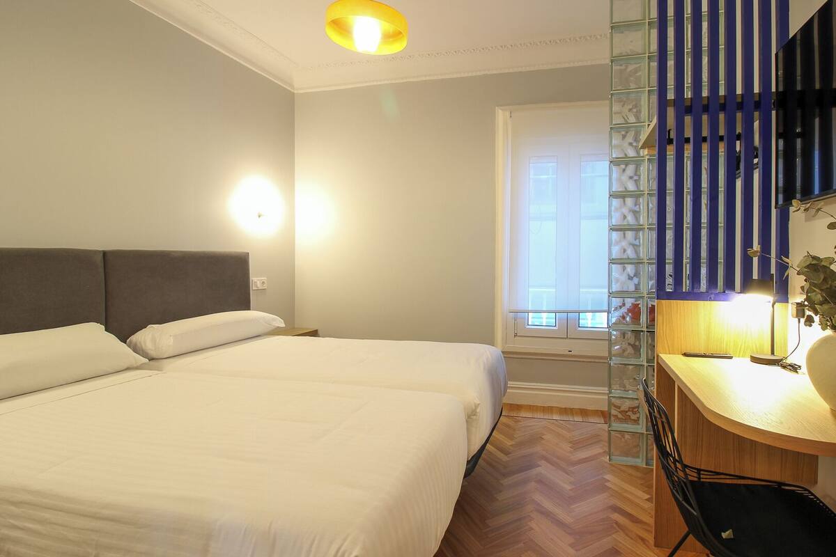 A hotel room with a bed, bedside table, lamp, and a view of a balcony with a railing.