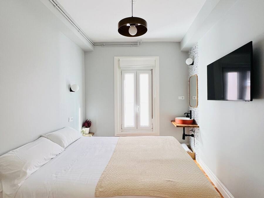 A bedroom with a bed, a bedside table, a mirror, a TV, and a window.