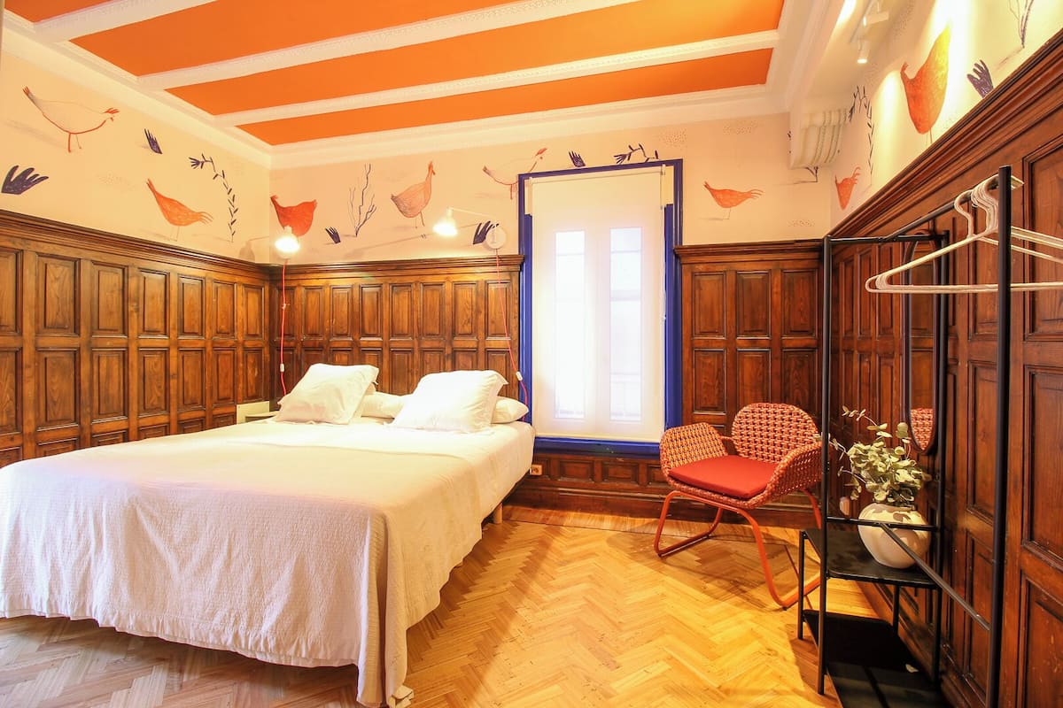 A bedroom with a large bed, wooden paneling, a window with curtains, and a chair with a vase.