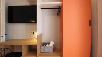 A compact room with a wooden desk, a black chair, a TV mounted on the wall, and a small shelf with a basket.