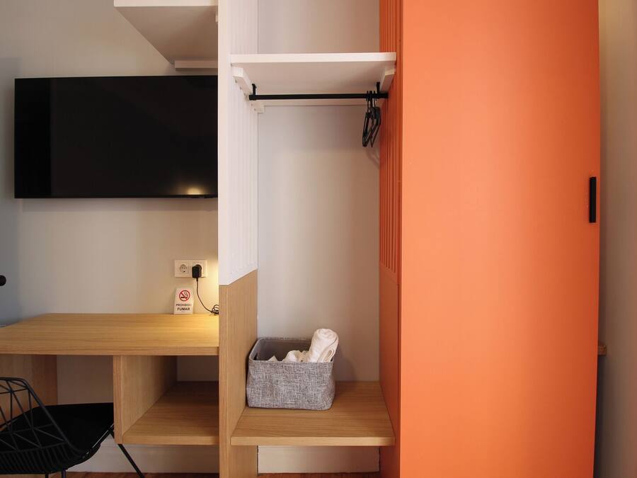 A compact room with a wooden desk, a black chair, a TV mounted on the wall, and a small shelf with a basket.