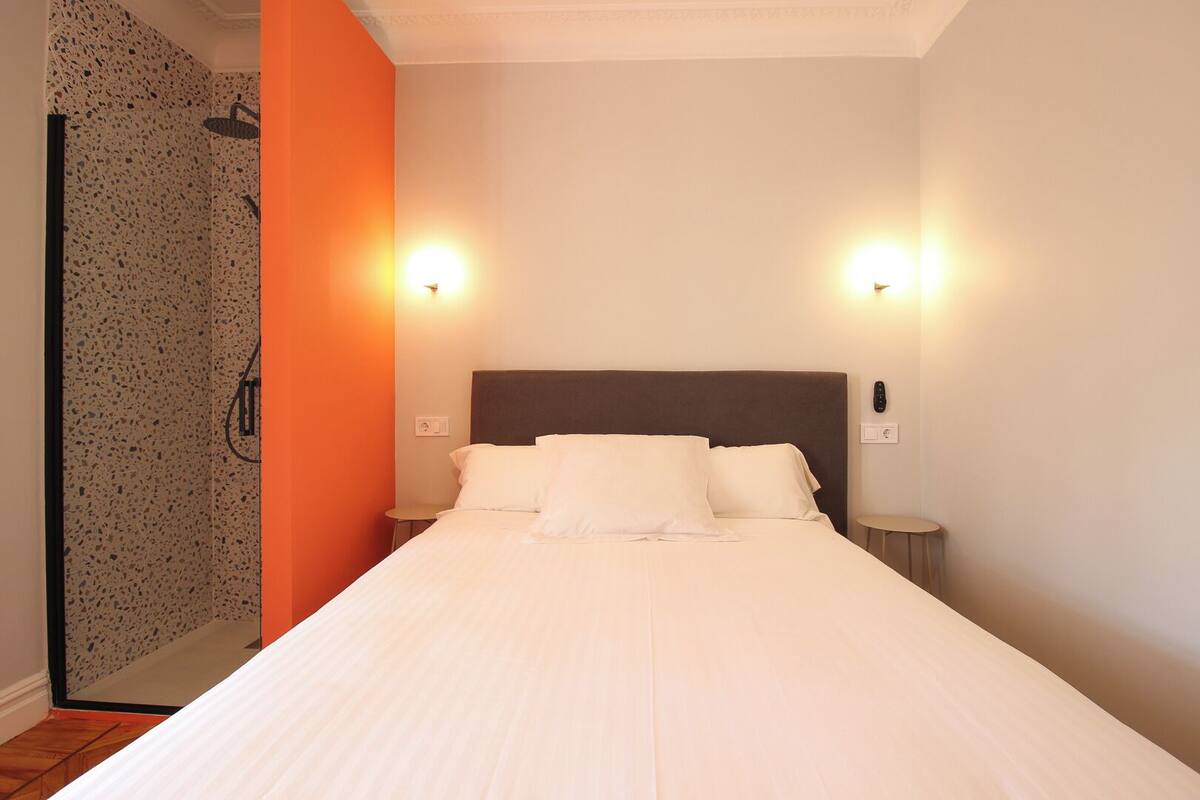 A hotel room with a bed, two bedside lamps, a shower area with a speckled wall, and a plain white wall.