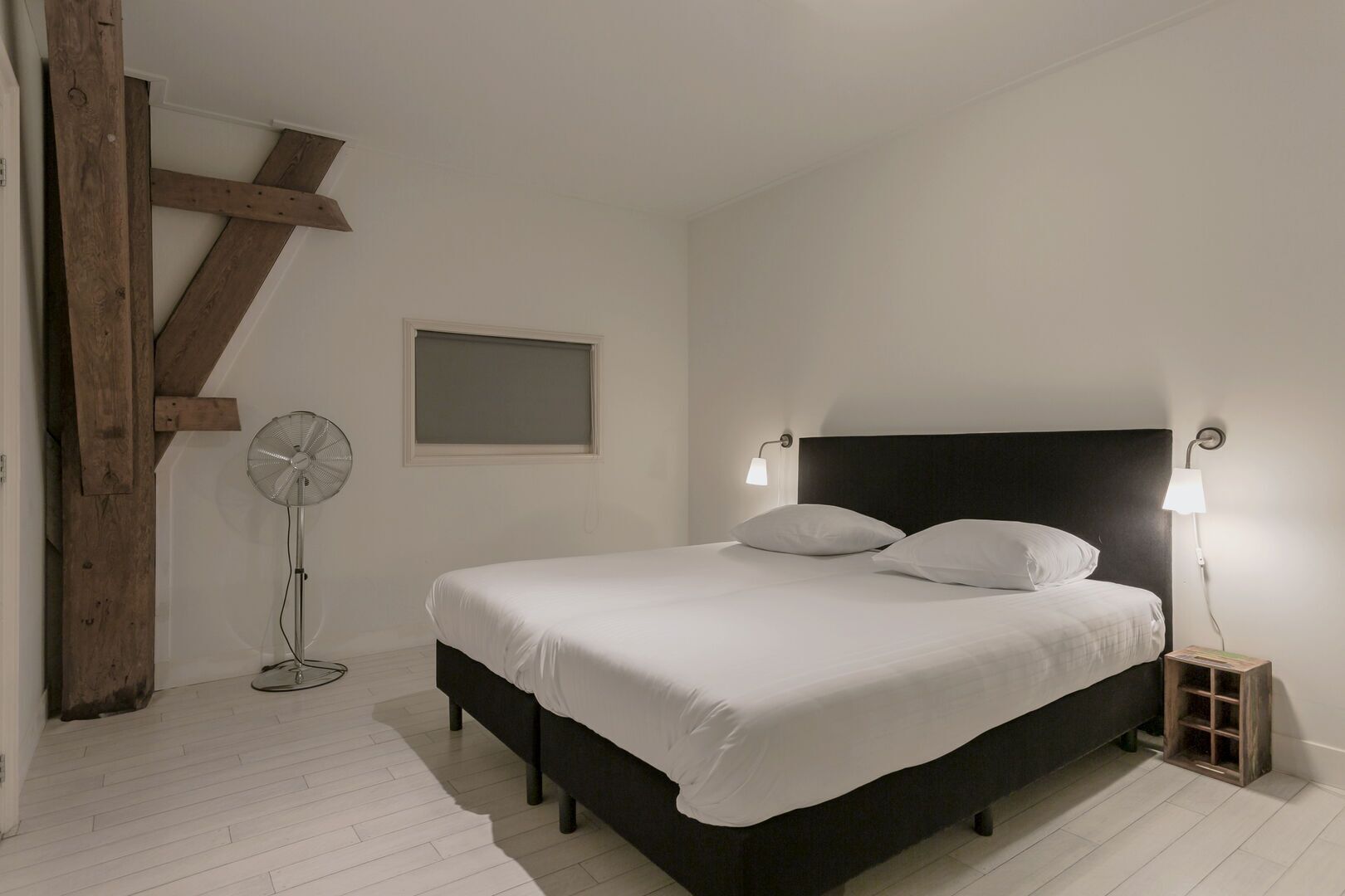 A bedroom with a bed, two bedside lamps, a standing fan, and a window.