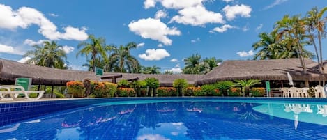 En klarblå pool omgiven av byggnader med halmtak, palmer och en klarblå himmel.