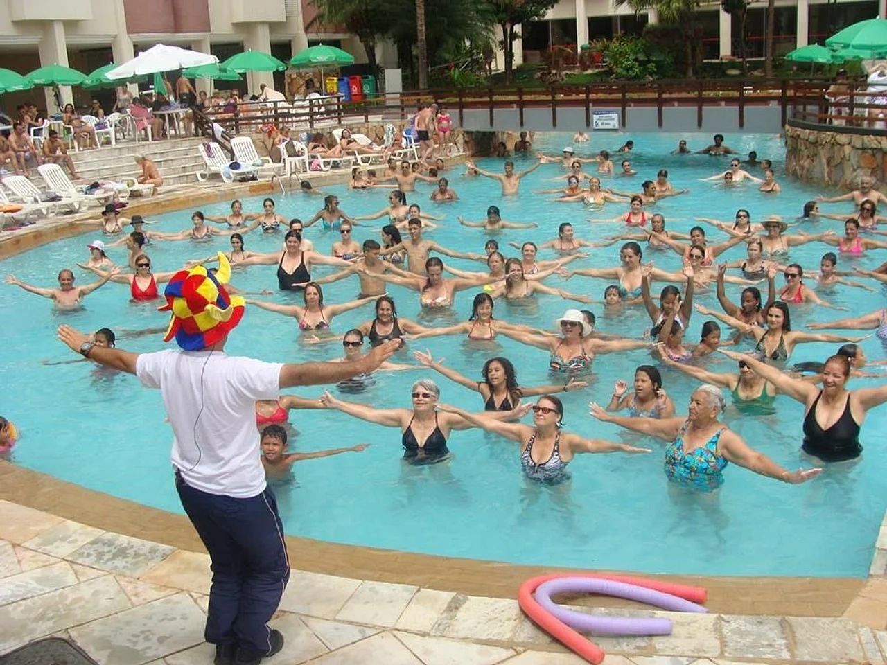 A swimming pool with people exercising, a person leading the exercise, and lounge chairs around the pool.