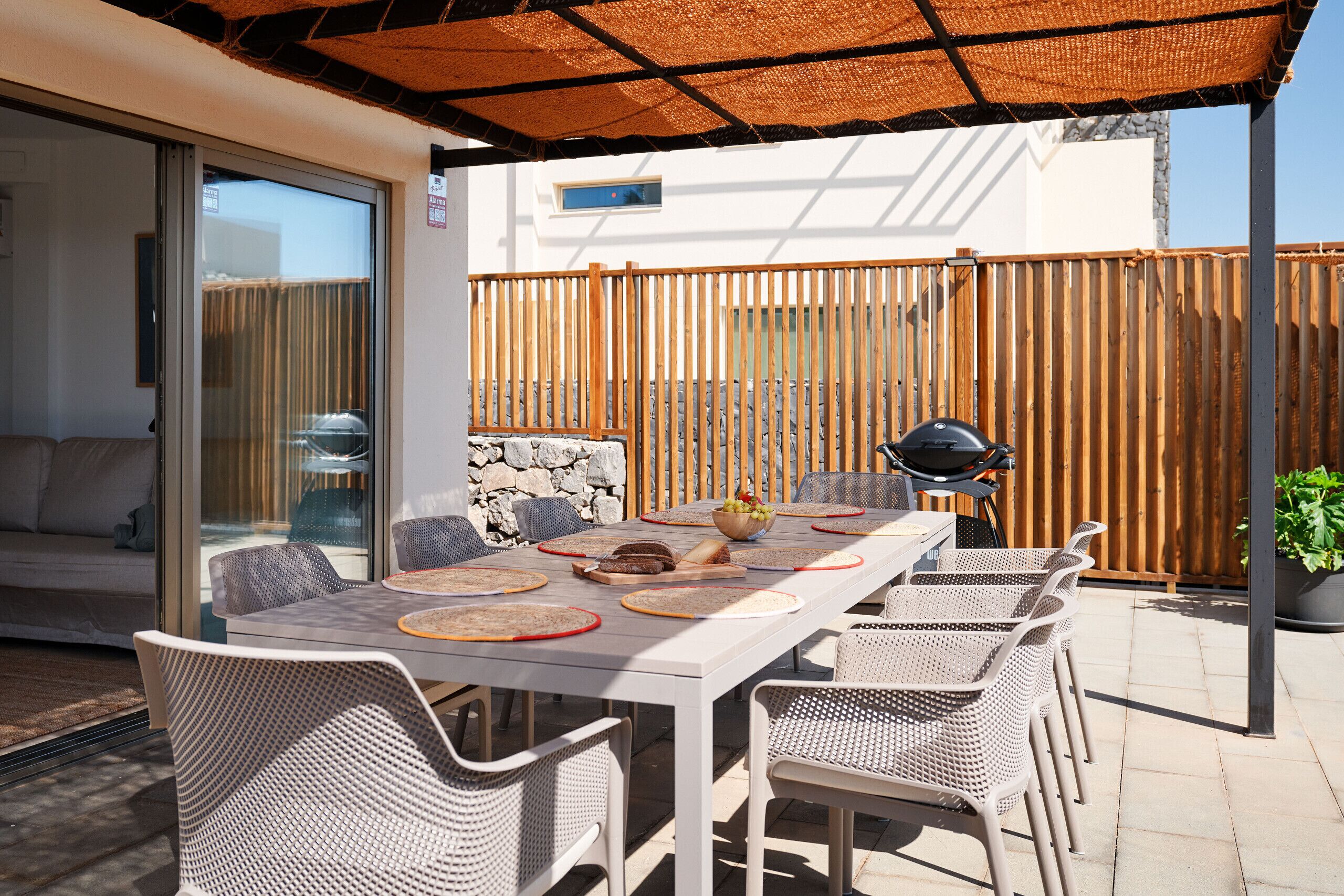An outdoor patio with a dining table set for four, surrounded by wicker chairs, a grill, and a wooden fence.