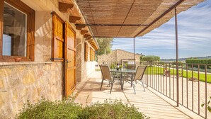 A covered patio with a table and chairs, stone walls, wooden doors, and a view of the countryside.