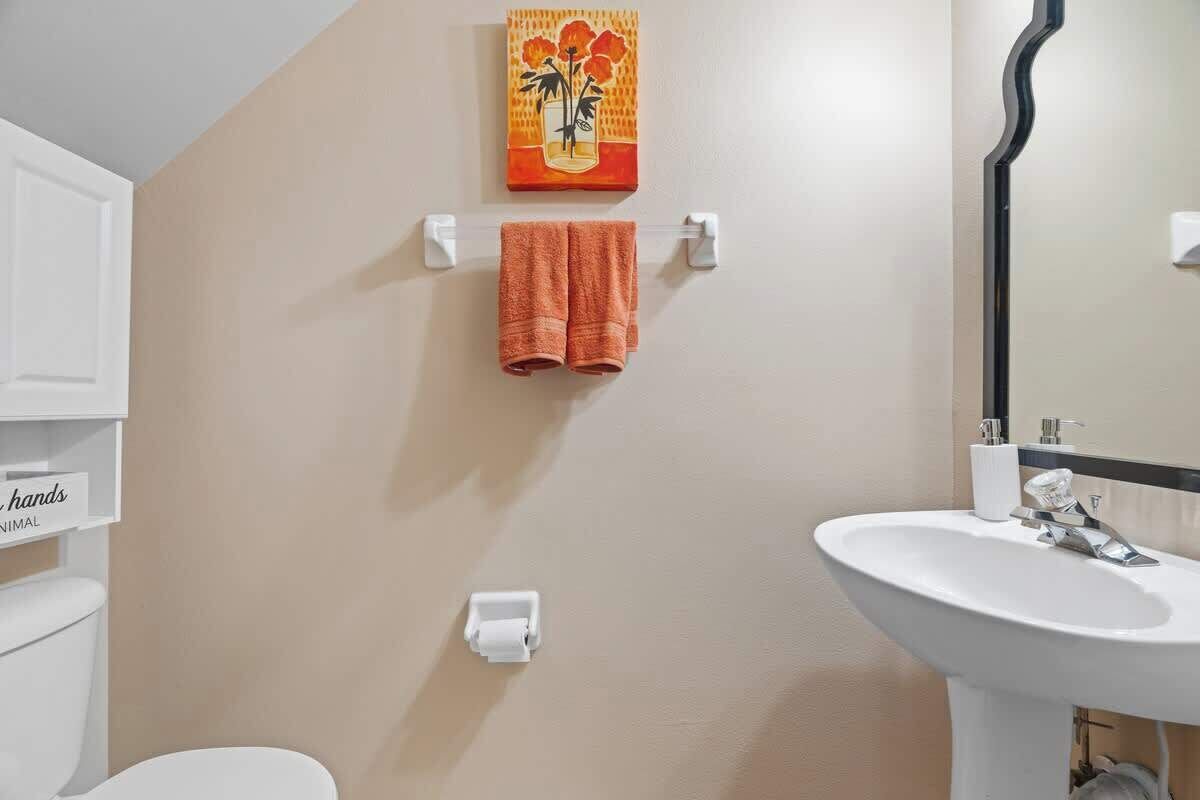A bathroom with a beige wall, a white sink, a mirror, a towel rack with an orange towel, and a toilet.