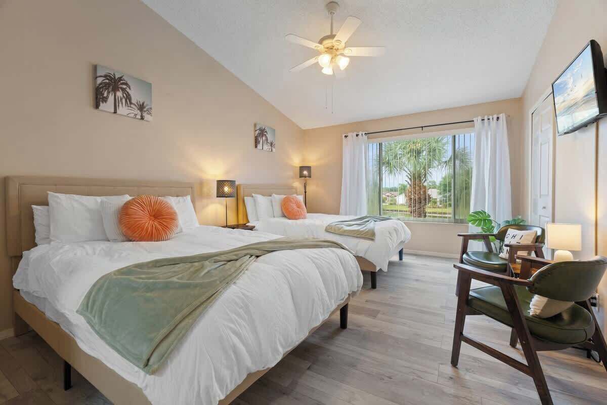 A hotel room with two beds, a ceiling fan, a desk with a chair, and a large window with curtains.
