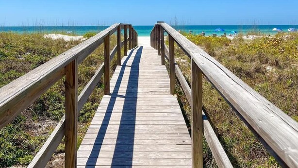 Une promenade en bois qui mène à une plage où l’eau est d’un bleu limpide et le ciel est dégagé.
