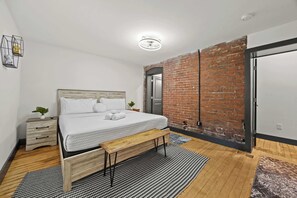 A bedroom with a wooden bed, a bench, a black and white rug, a brick wall, and a door. - NEW Cozy Home in Shelby Park Near Cafes & Stores (Louisville)