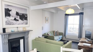 A living room with a fireplace, green sofa, white chairs, a television, and a framed photograph on the wall.