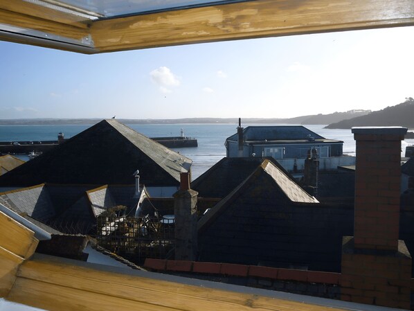 A coastal view from a building with a wooden roof and brick chimney, overlooking a harbor with a lighthouse and a pier.