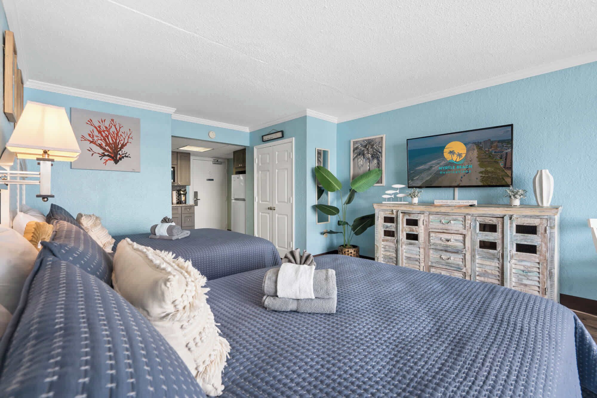 A hotel room with two beds, a TV, a kitchenette, and a view of the beach.