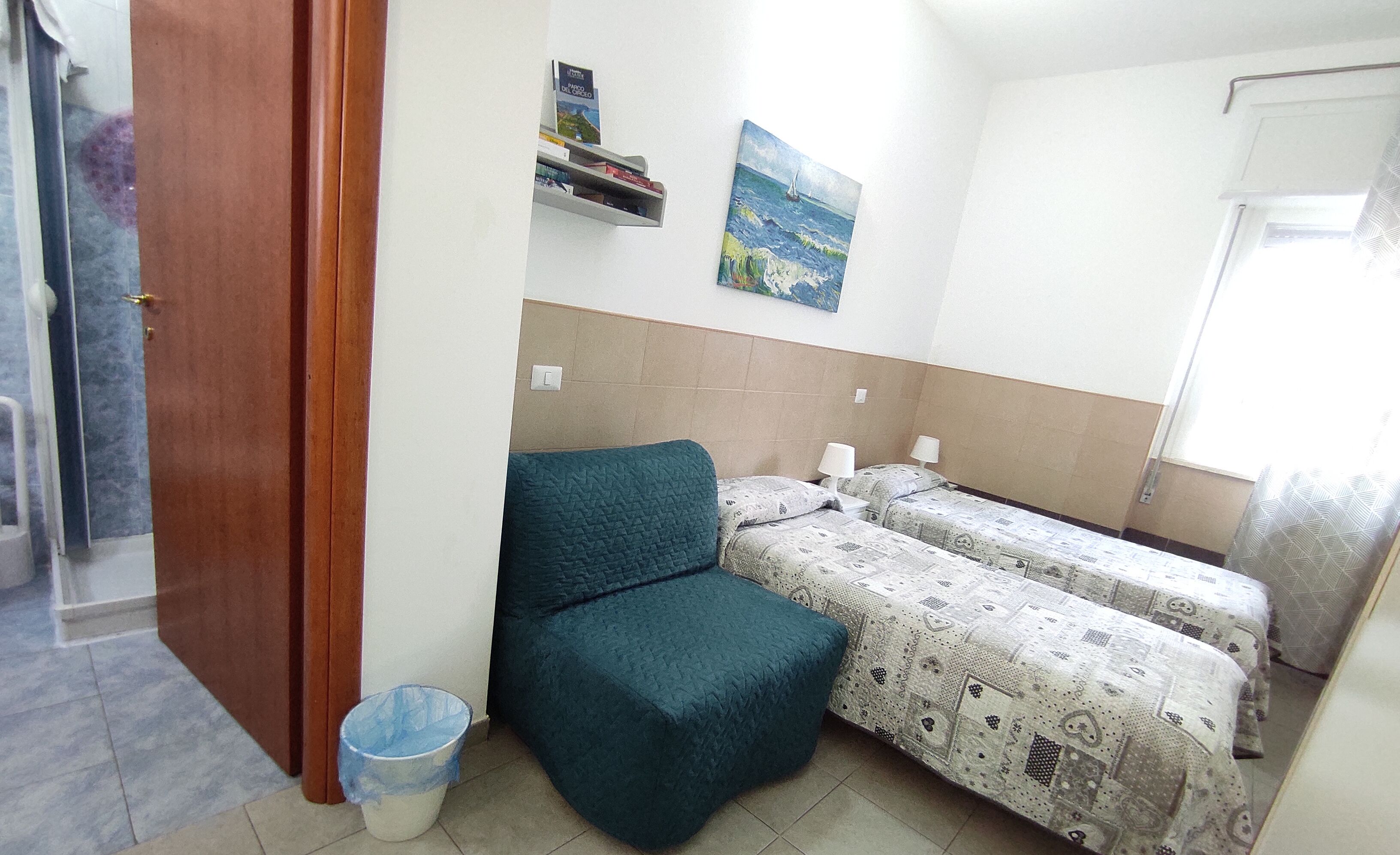 A small, simple room with two single beds, a small chair, a trash bin, a shelf with books, and a painting on the wall.