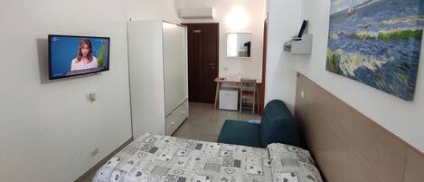 A hotel room with a bed, a desk with a chair, a small refrigerator, a TV, and a painting on the wall.