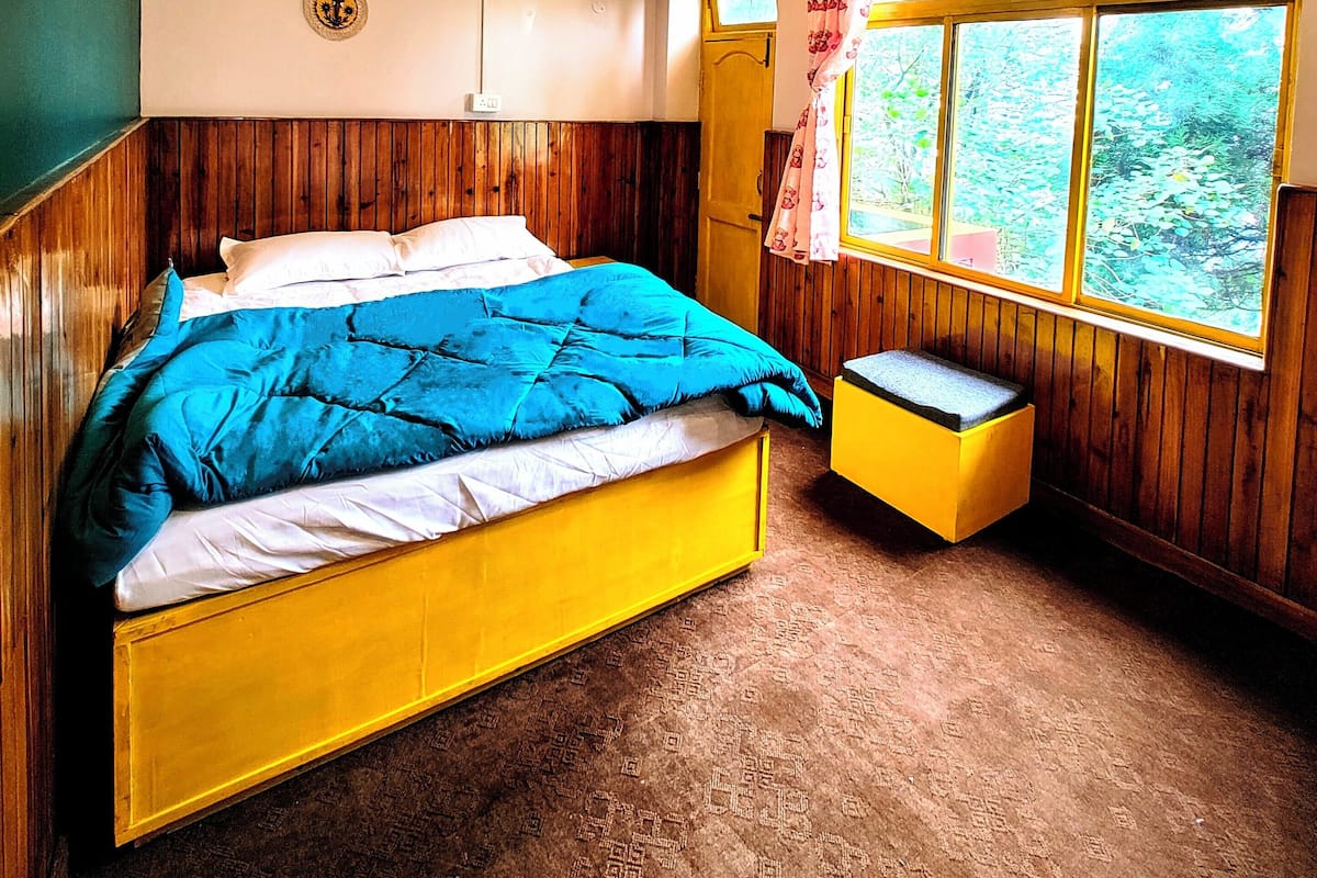 A room with a bed, a window with curtains, a wooden bench, and a wall-mounted light fixture.