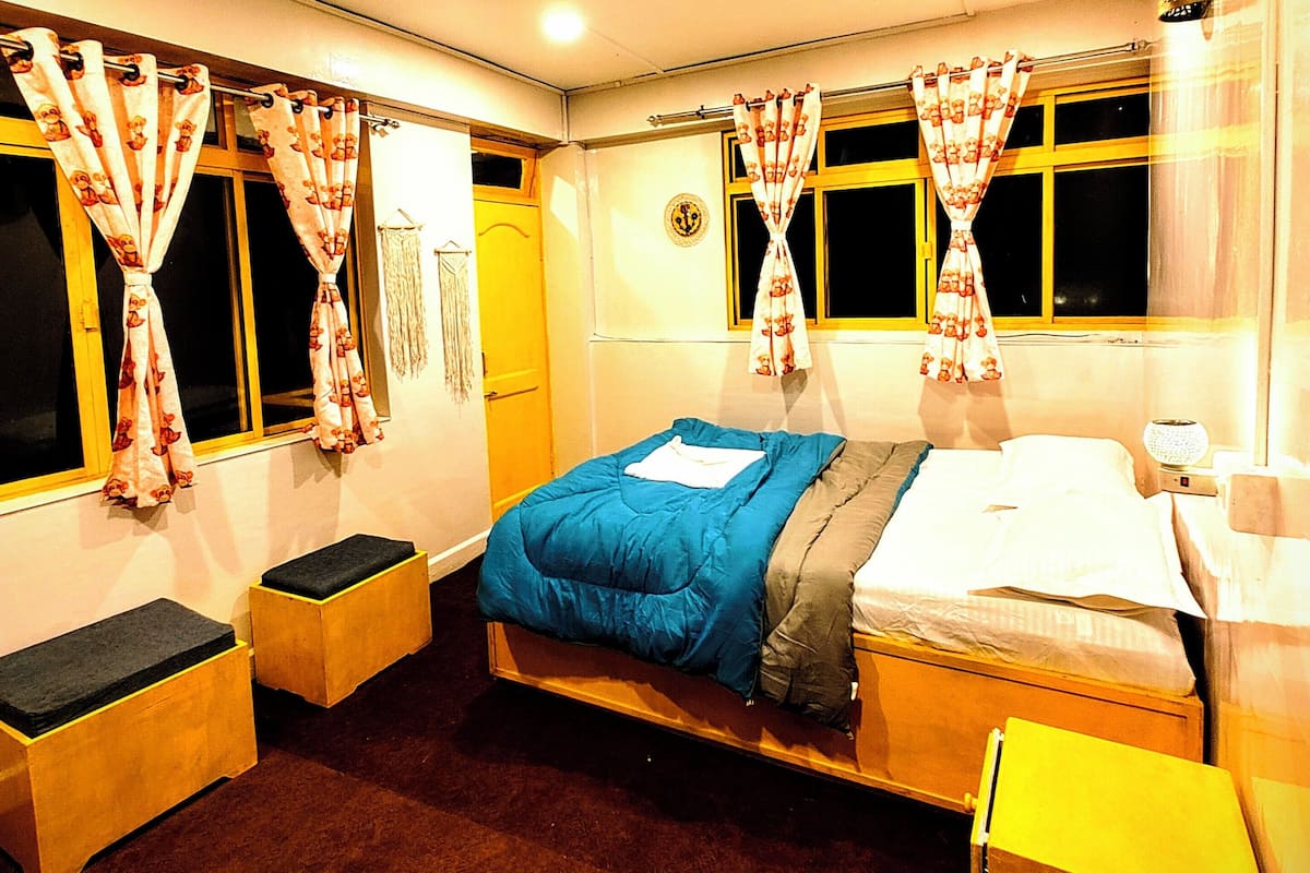A room with a bed, two bedside tables, a window with curtains, a wall clock, and a small table with a lamp.