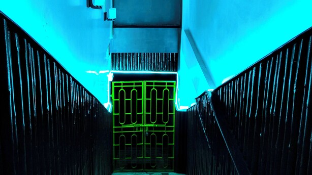 Un couloir étroit avec un escalier menant à un palier. Les murs sont peints dans une couleur claire, et il y a une fenêtre à carreaux sur un des côtés.