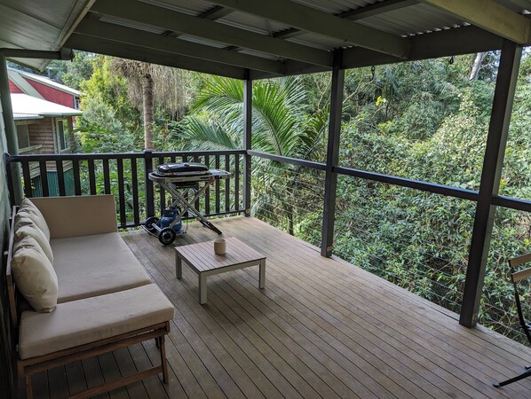A covered outdoor area with a wooden deck, a beige sofa, a small table, and a grill cart.