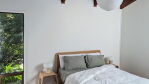 Un dormitorio con techo de madera, una cama con ropa de cama blanca, una mesita de noche, una lámpara de noche y una ventana con vistas a zonas verdes.