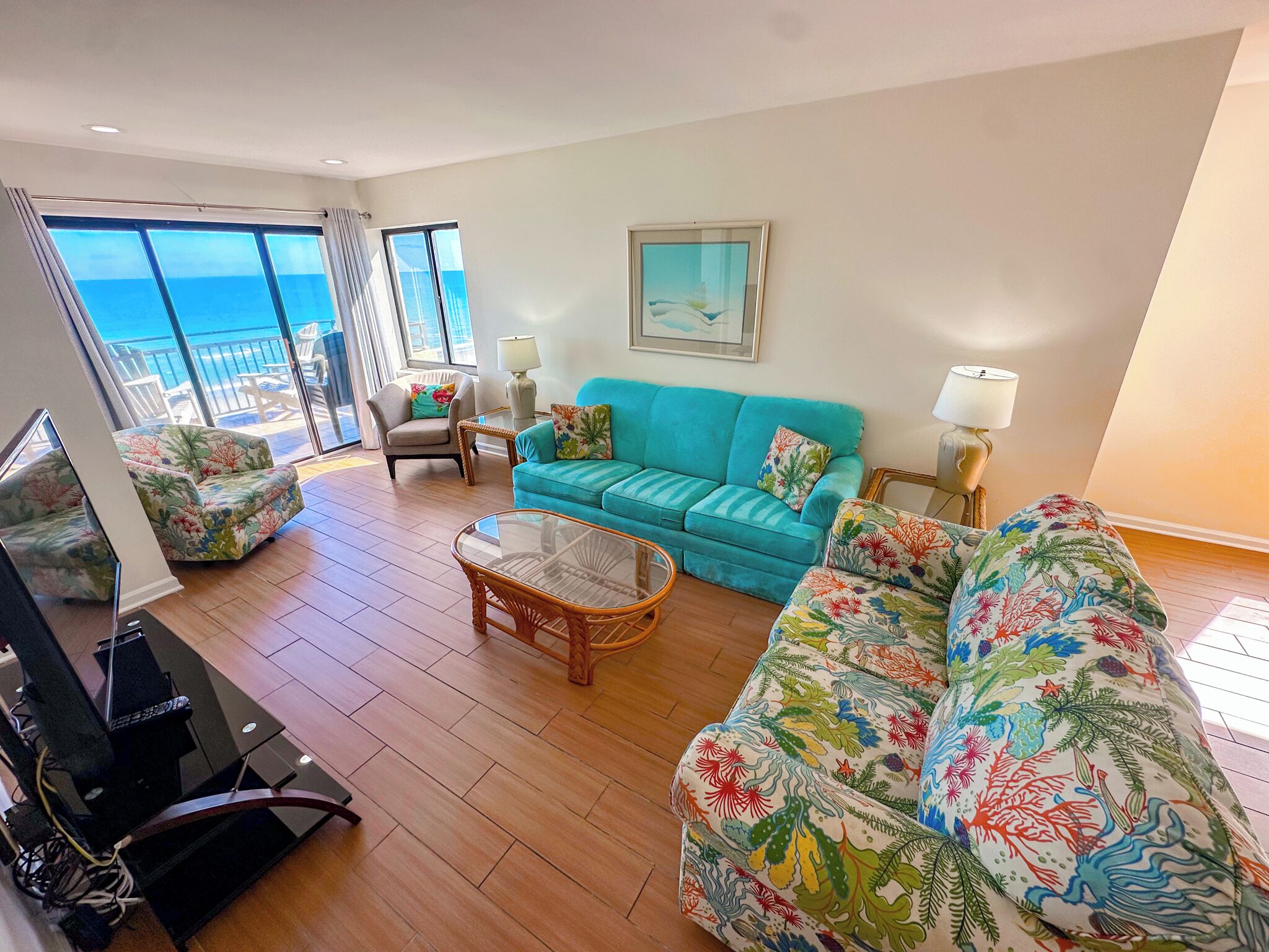 Ein geräumiges Wohnzimmer mit einem türkisfarbenen Sofa, floralen Armlehnstühlen, einem Glastisch und einem Fernseher. Große Fenster bieten Blick auf den Ozean.