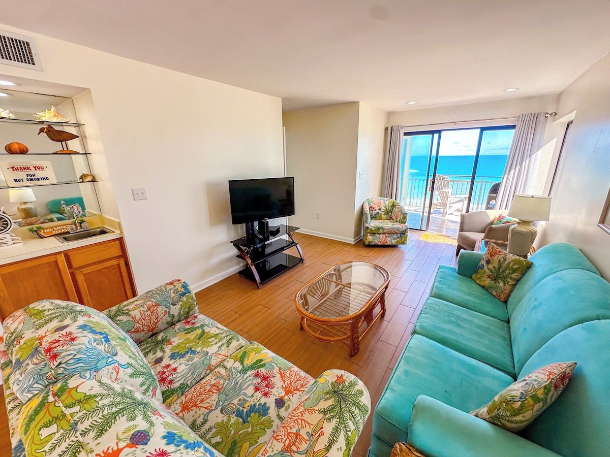 A living room with a sofa, a television, a wicker chair, and a view of the ocean through large windows.