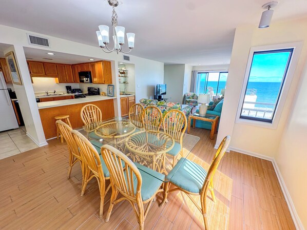 Un espacio habitable amplio con una mesa de comedor de vidrio rodeada de sillas de mimbre, una cocina con armarios de madera y un gran ventanal con vistas al océano.