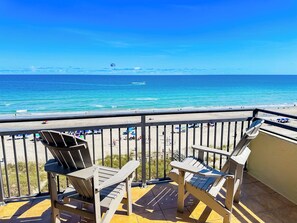 A balcony with two Adirondack chairs overlooking a beach with people and umbrellas.