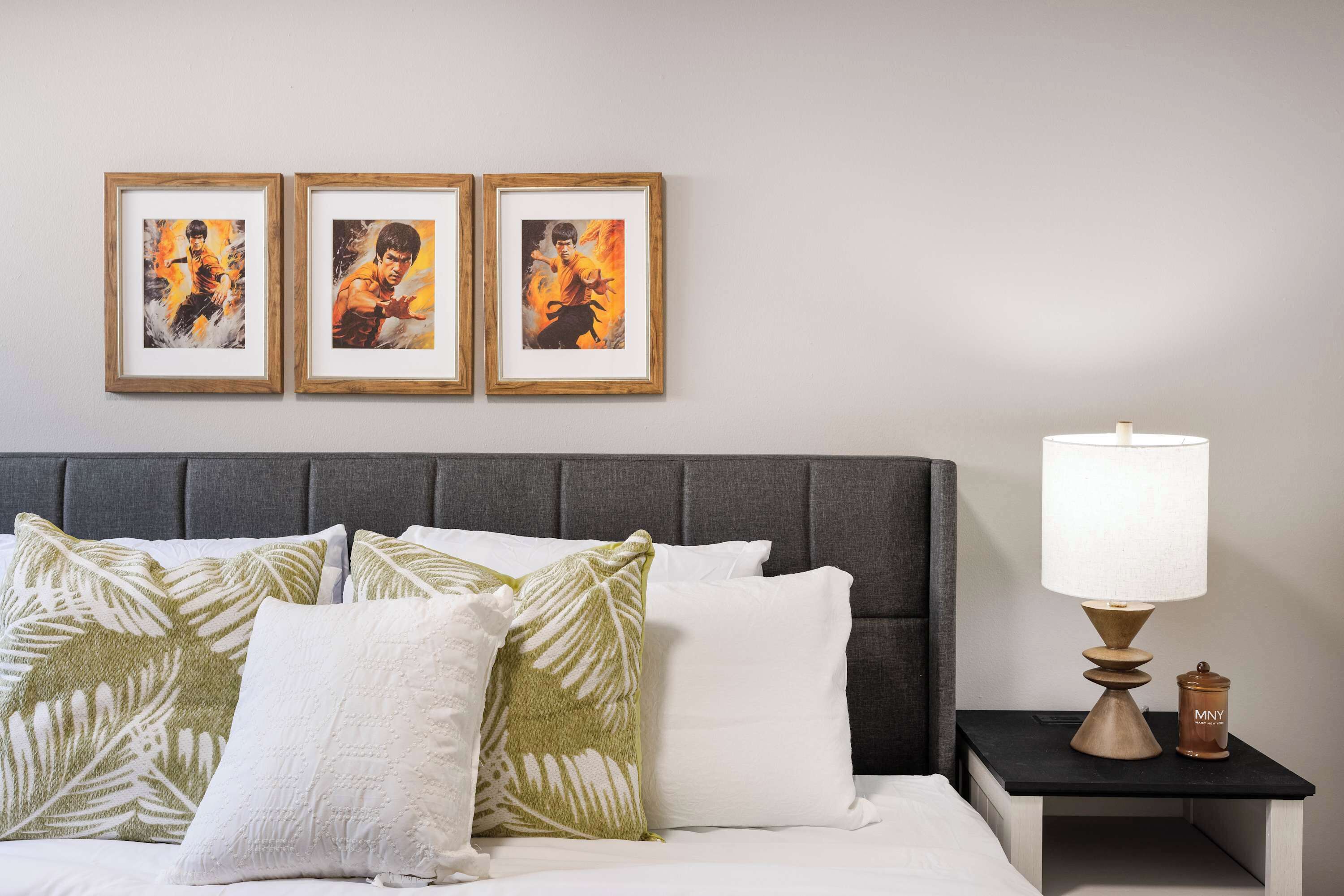 Un lit bien fait, avec des oreillers blancs et verts, une tête de lit rembourrée de couleur grise et trois tableaux encadrés accrochés au mur.