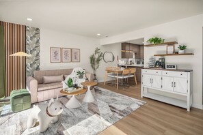 A modern living room with a sofa, coffee table, and dining area. There is a kitchen with white cabinets and a coffee machine.