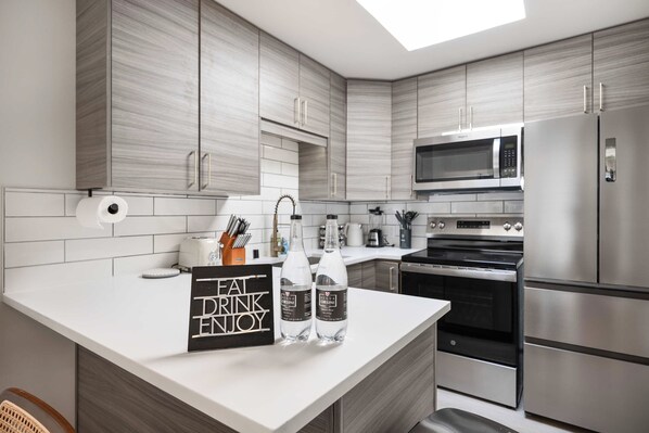 A modern kitchen with grey cabinetry, a white countertop, a built-in microwave, a stainless steel refrigerator, and a sign that reads "Eat Drink Enjoy".