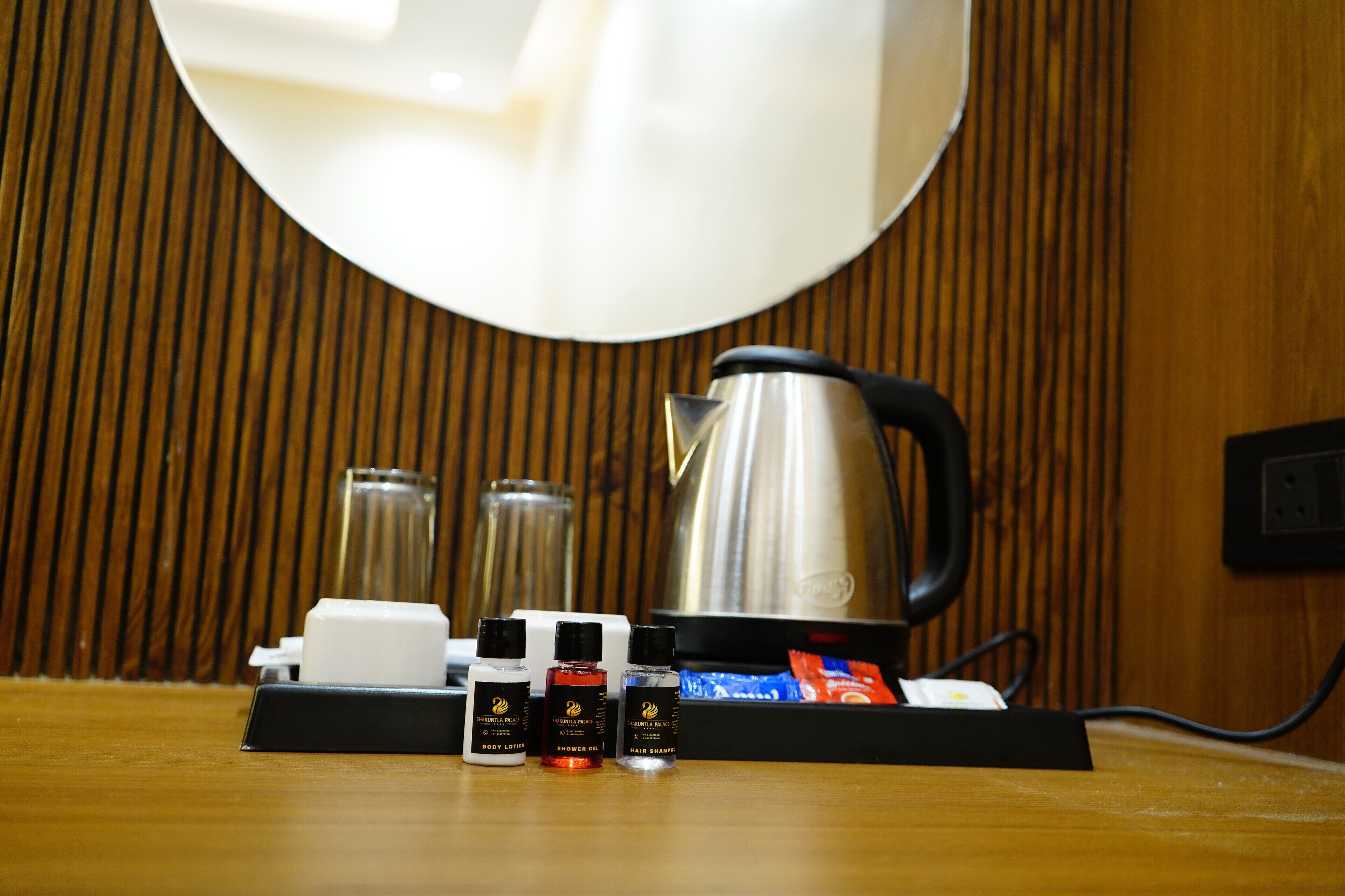 Une table en bois sur laquelle se trouve un plateau noir, et sur ce plateau, une machine à café, un verre et trois flacons de produits de toilette.