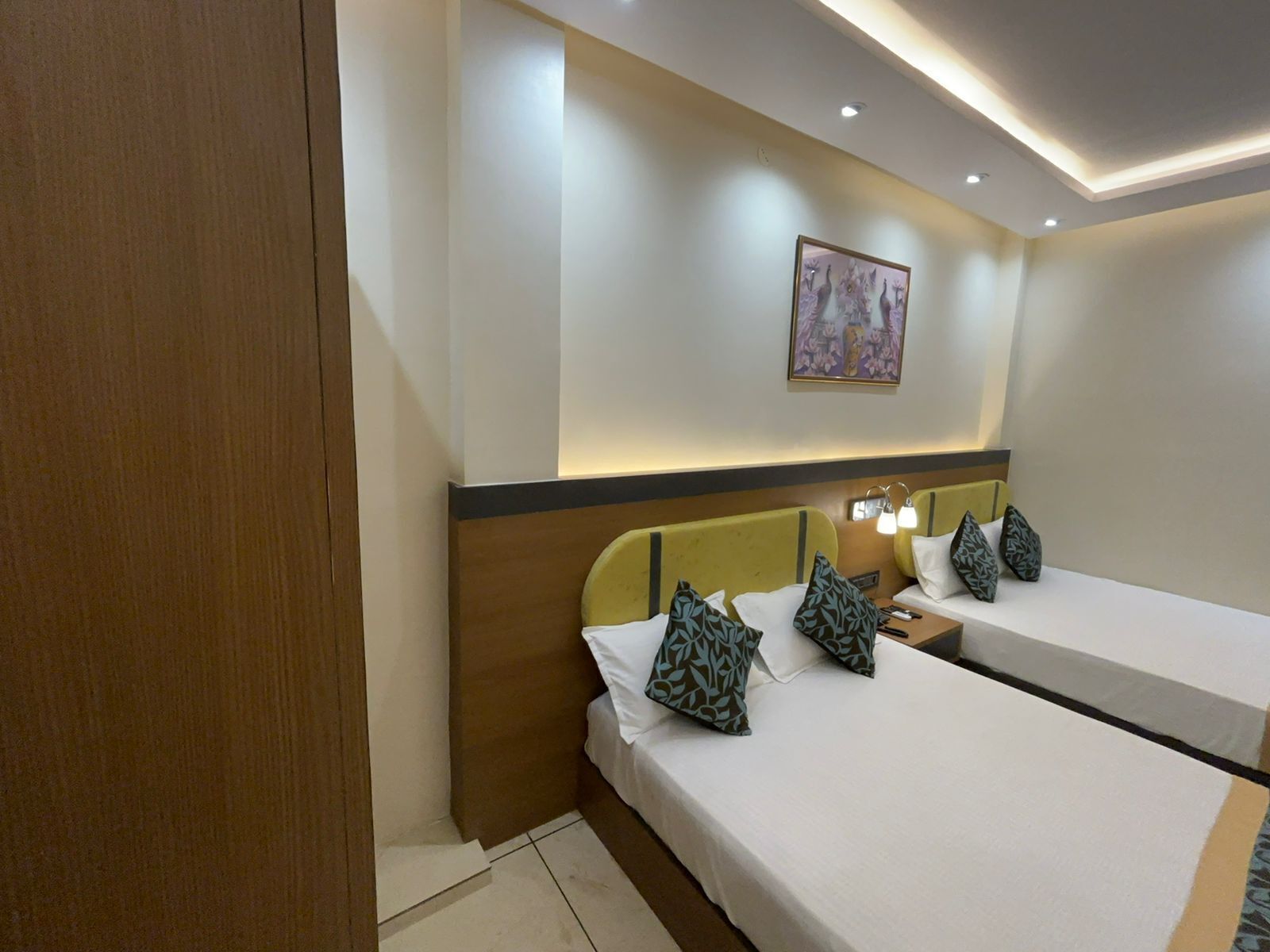 A hotel room with two beds, a wooden wardrobe, a nightstand with a lamp, and a painting on the wall.