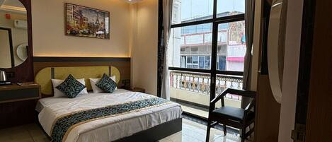 A modern hotel room with a large bed, a ceiling fan, a window with a view of the city, and a small table with a chair.