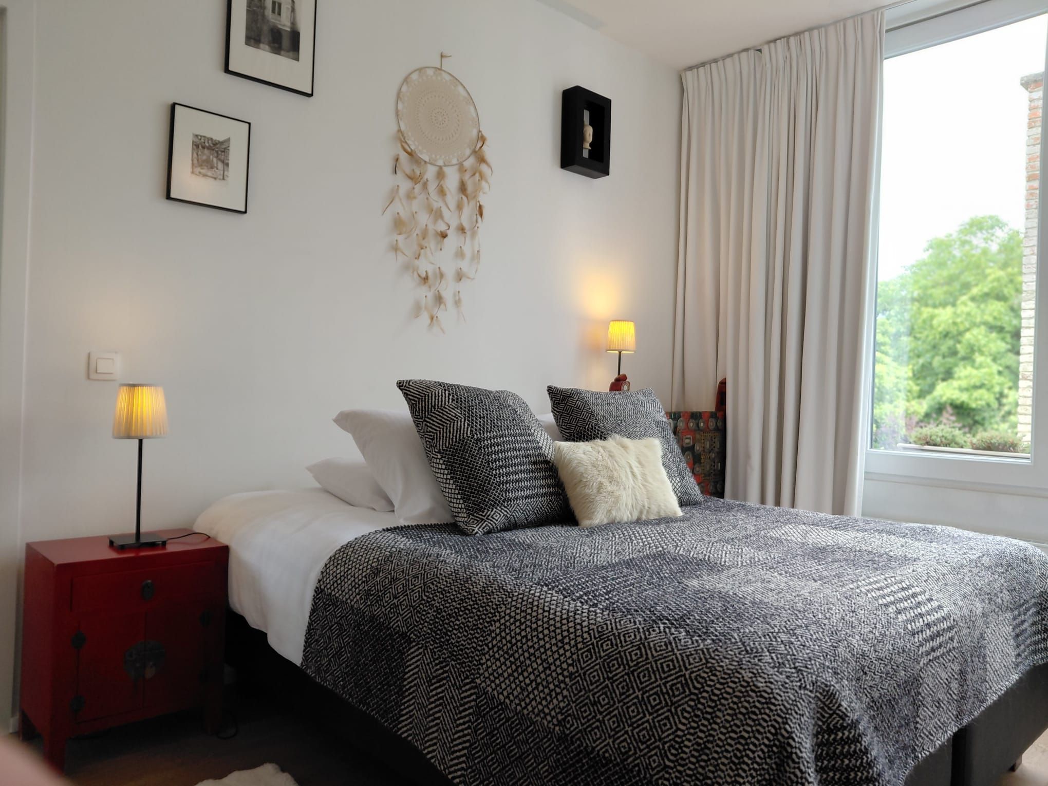 Une chambre à coucher avec un lit, deux lampes de chevet, une table de nuit rouge, un attrape-rêves fixé au mur et une fenêtre avec des rideaux.